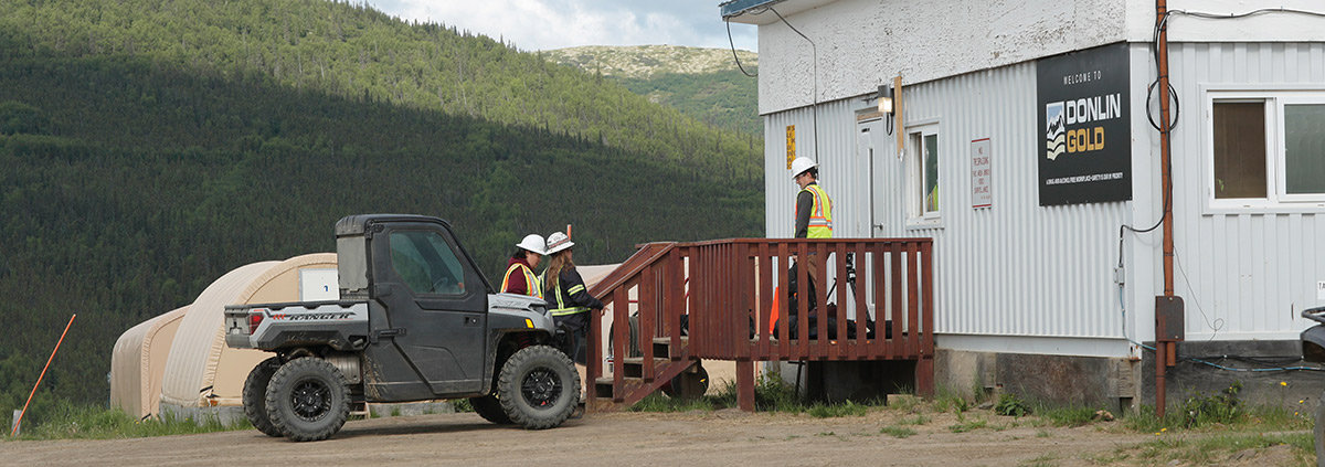 Workers stand near a Donlin Gold office building and an all-terrain vehicle in a mountainous area.