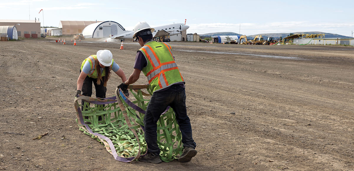 Two workers in safety vests handle a large green cargo net on a dirt airfield.