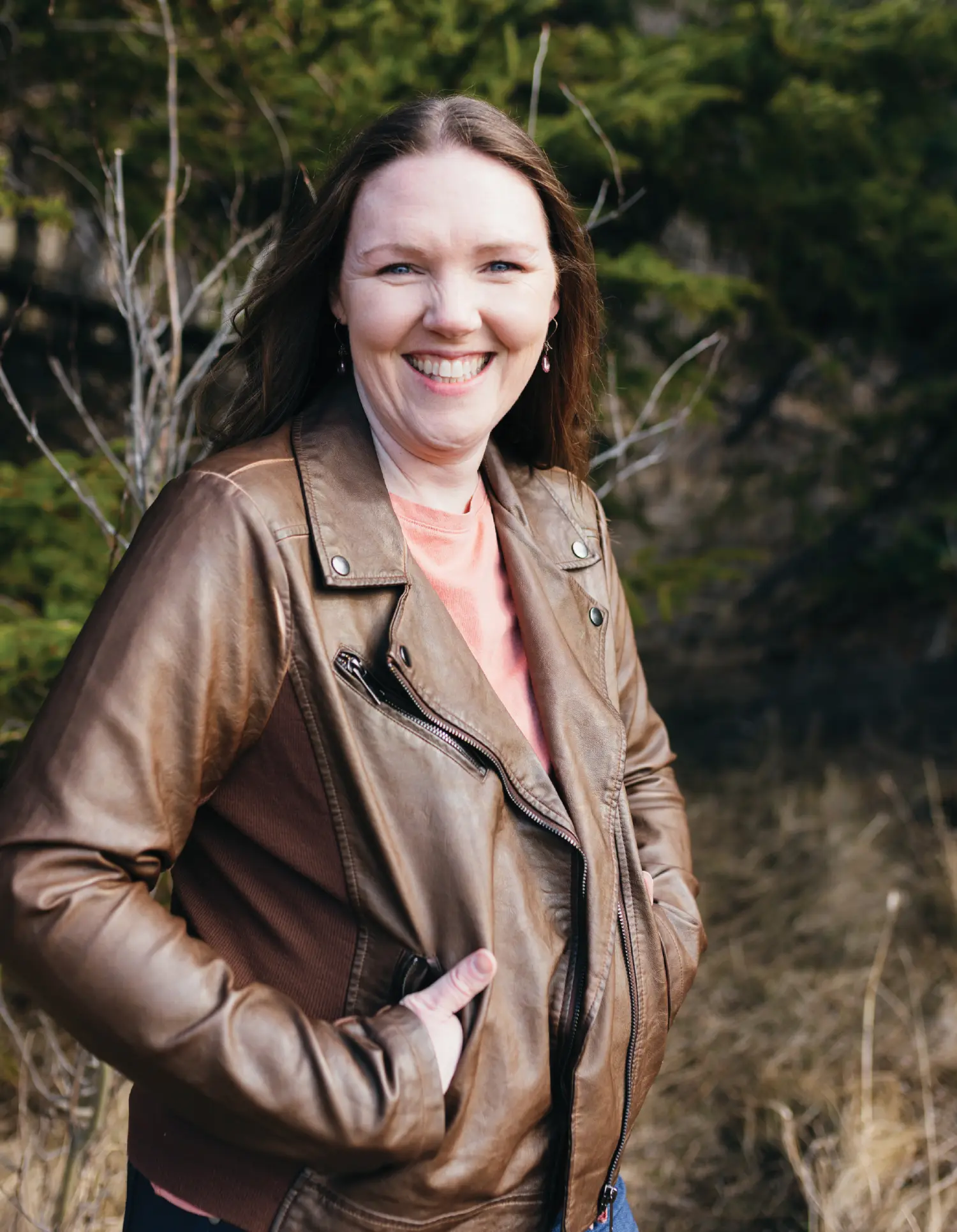 Sasha Swift, a smiling woman in a brown faux-leather moto-style jacket over a peach t-shirt with her hands resting in her jacket pockets standing outside in nature