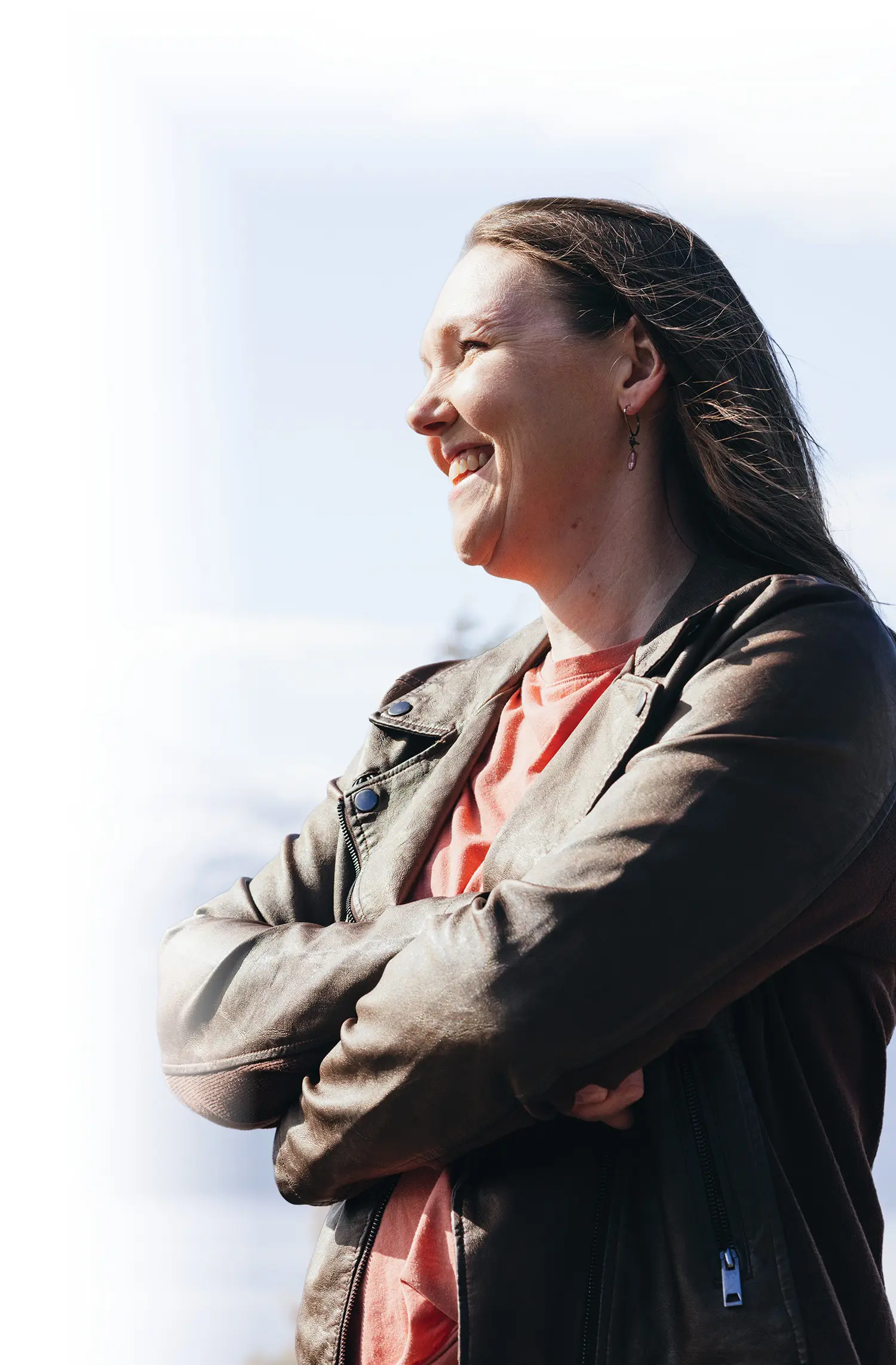 Sasha Swift, a smiling woman looking away off into the far distance as she has her arms crossed wearing a brown faux-leather moto-style jacket over a peach t-shirt