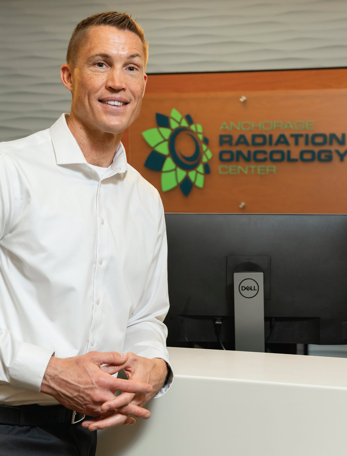 A smiling man in a white button-down shirt stands by a reception desk in front of an Anchorage Radiation Oncology Center sign.