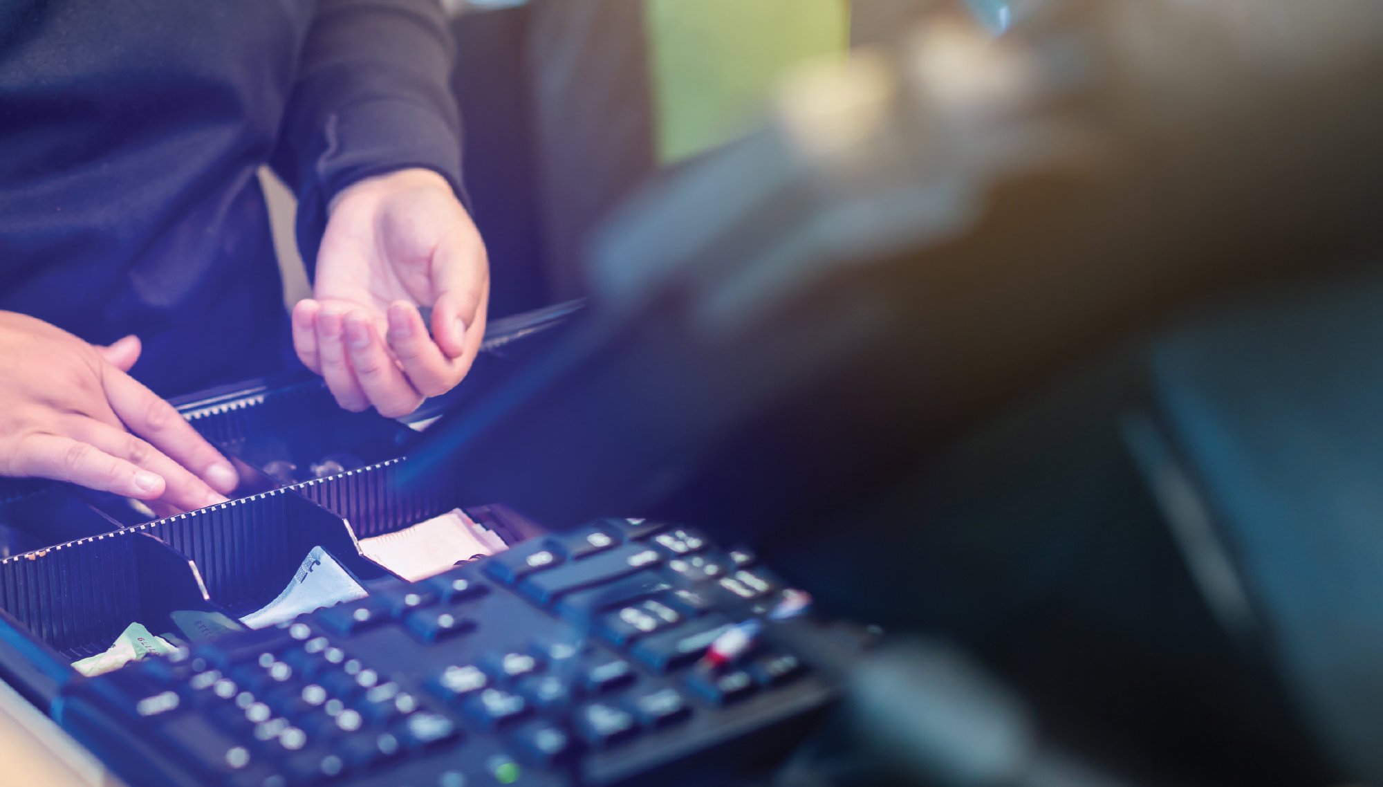 Close-up of a cashier's hands reaching into an open cash register drawer to count out change at a checkout counter.