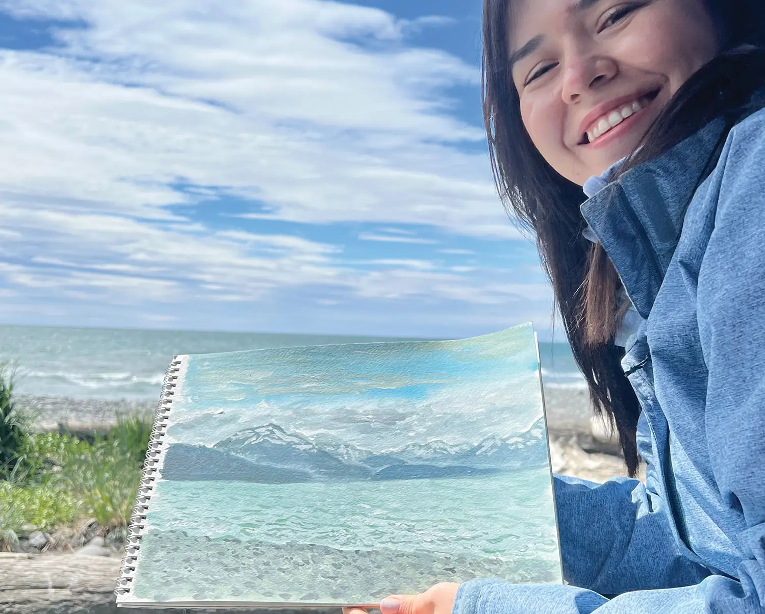 A smiling woman holds a spiral sketchbook displaying a landscape painting of mountains and water, set against a real background of a sea, rocky beach, and cloudy blue sky.