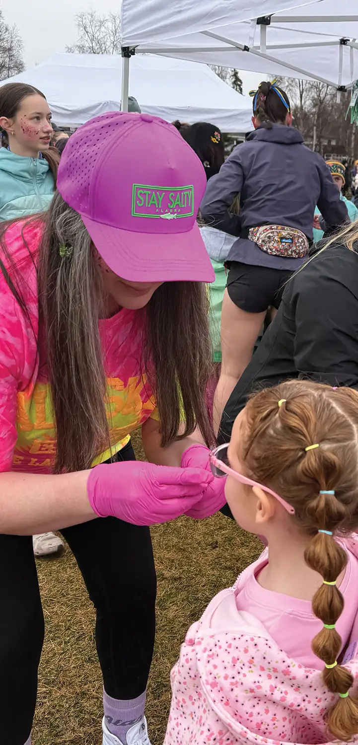 A person wearing a pink "Stay Salty Alaska" cap and pink gloves leans down to interact with a young girl wearing glasses and braided hair at an outdoor event.