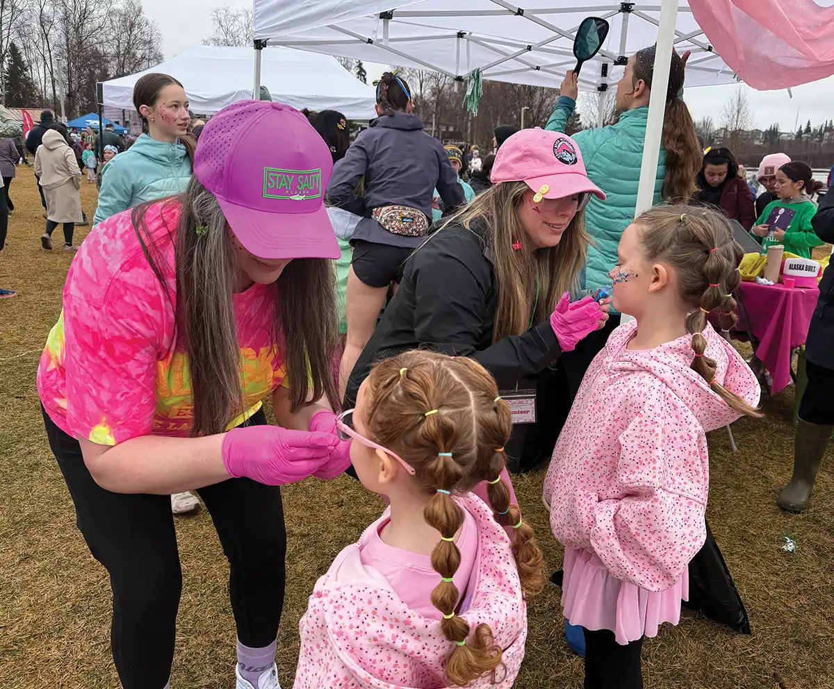 Two volunteers in pink hats and gloves apply face paint and glitter to young girls with braided hair at an outdoor community event with white tents.