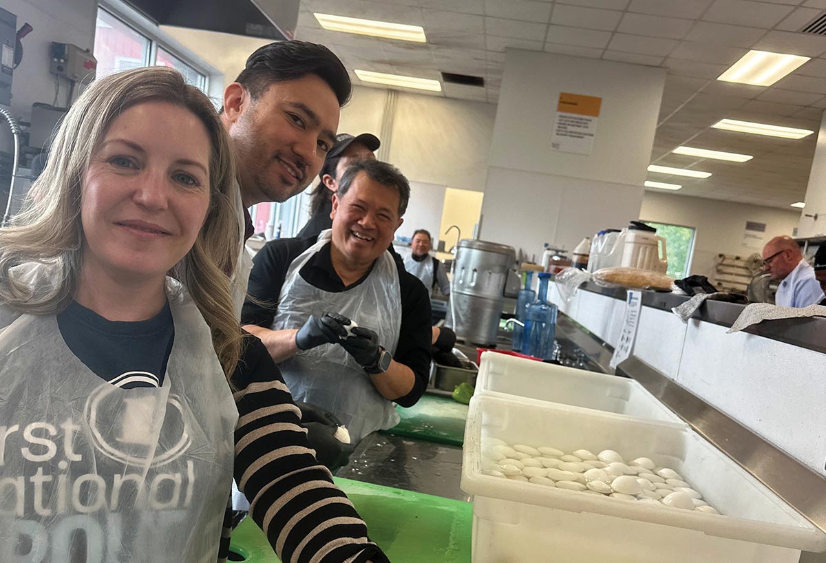 A group of smiling volunteers wearing clear plastic aprons prepare food inside a commercial kitchen, handling hard-boiled eggs next to large white storage bins.