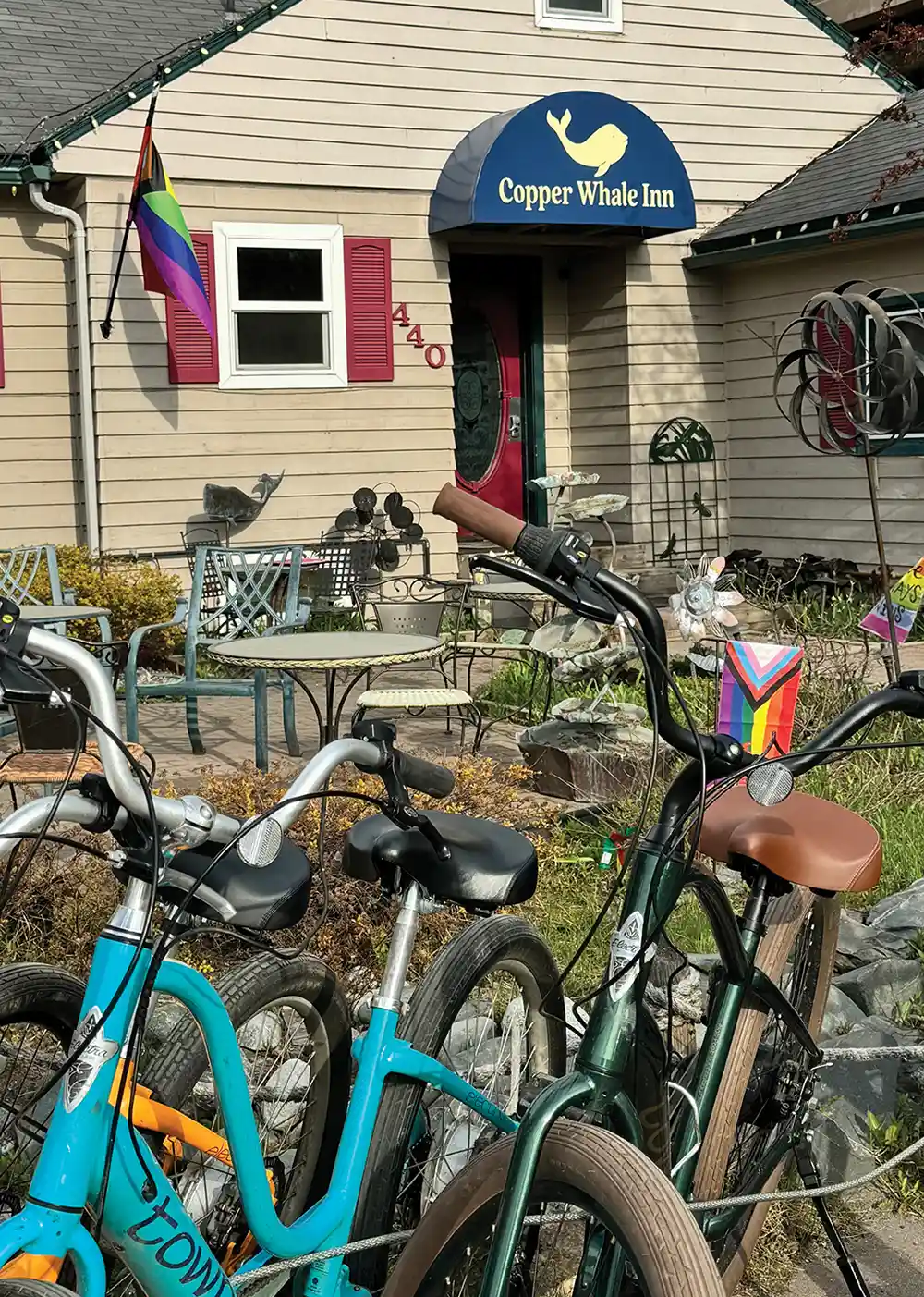 Bicycles are parked in the foreground outside the Copper Whale Inn, which features a blue awning with a whale logo, red shutters, an outdoor patio, and displayed Pride flags.