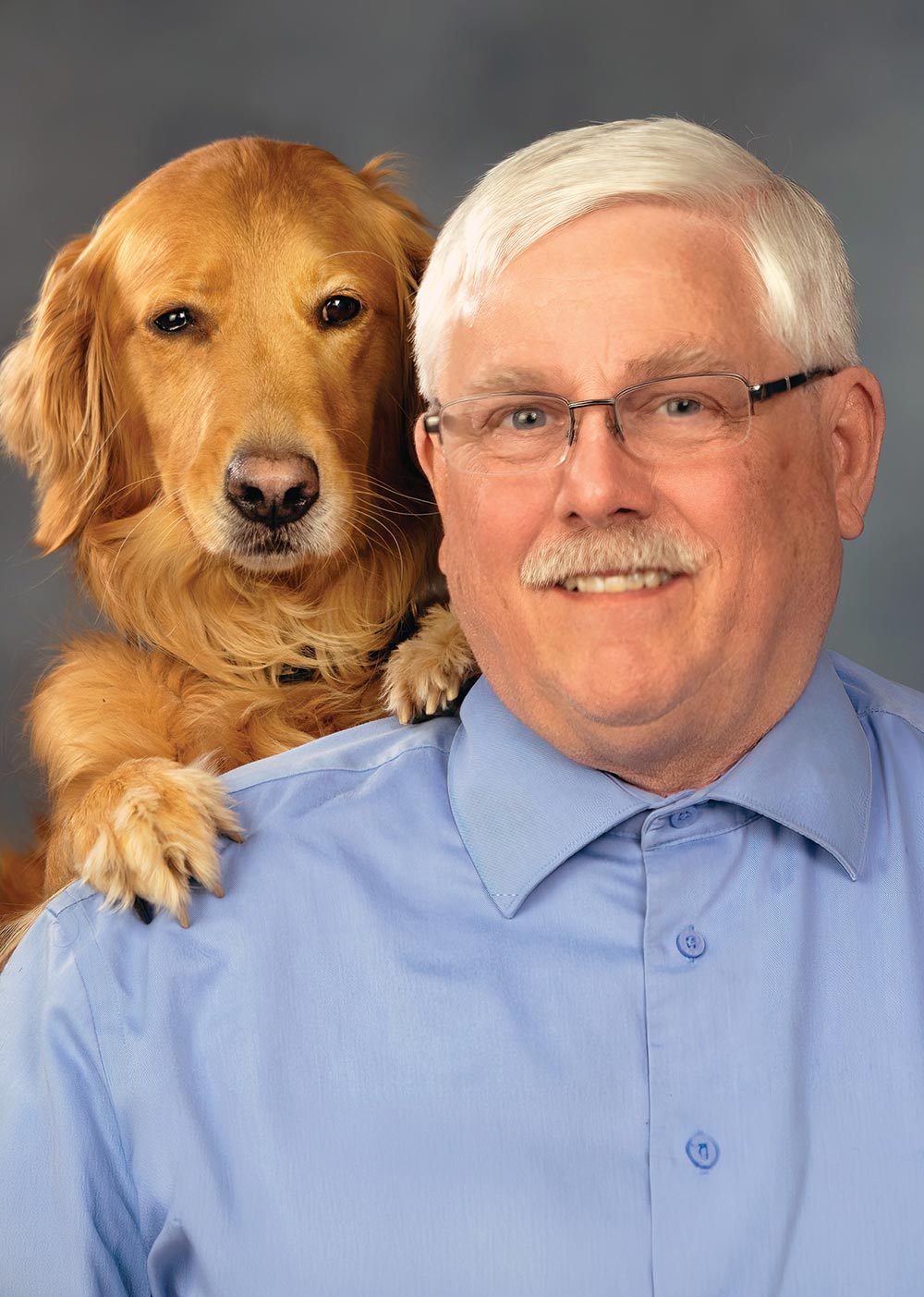A studio portrait of a smiling older man with glasses and a mustache wearing a blue shirt, with a golden retriever resting its paws on his shoulder against a gray background.