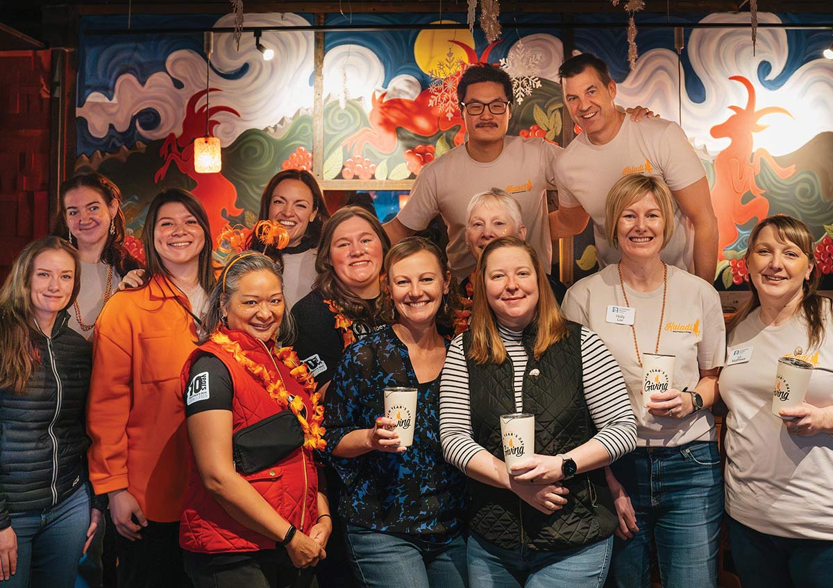 A large group of smiling people pose together in front of a colorful stylized mural, with several individuals holding white tumblers that read "New Year's Day Giving."