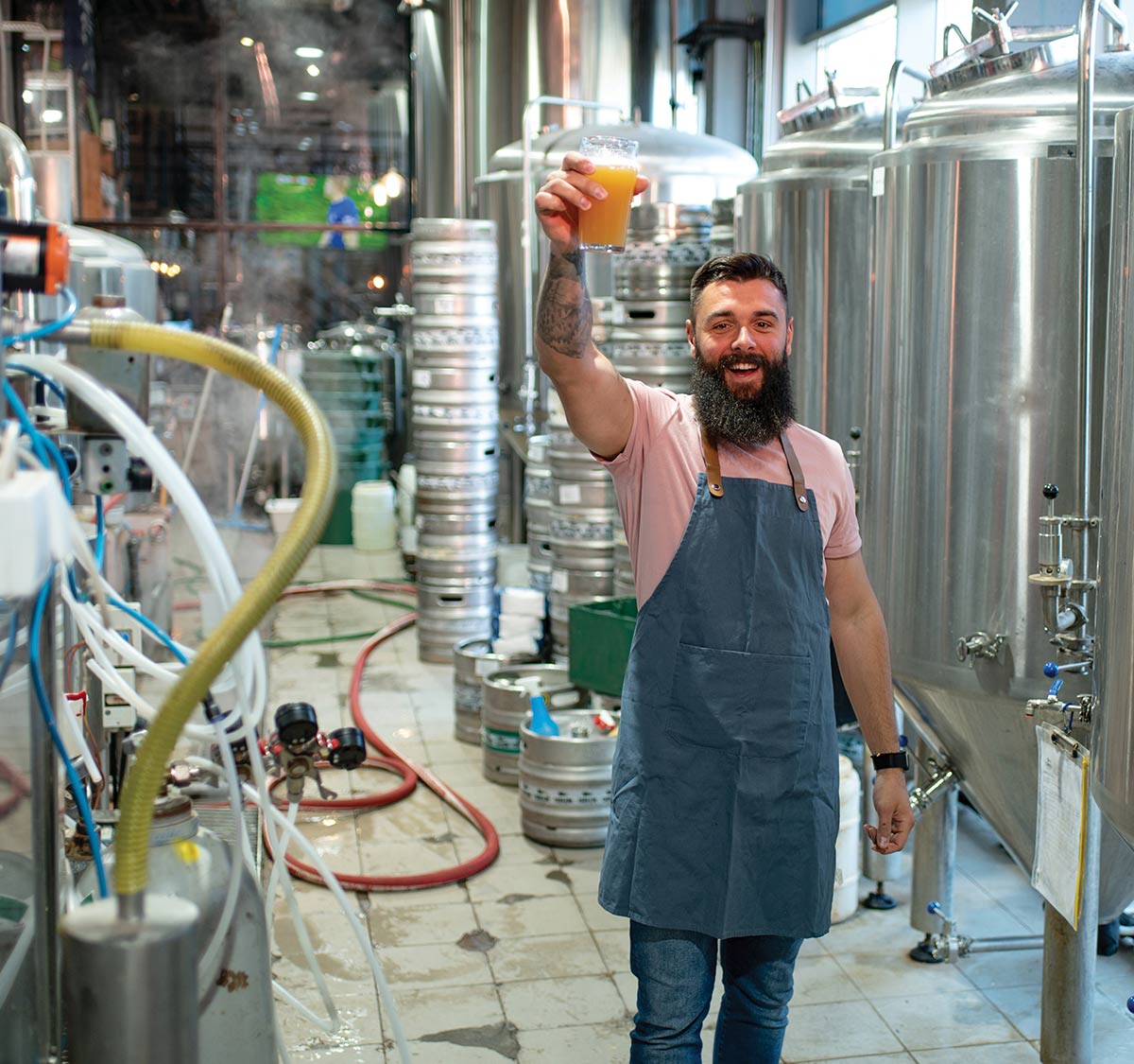 A smiling, bearded man in a blue apron raises a glass of beer inside a brewery surrounded by large stainless steel tanks and stacked metal kegs.