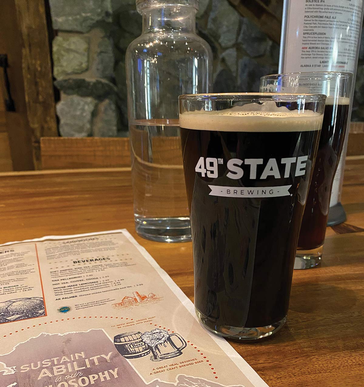 A pint glass filled with dark beer featuring the "49th State Brewing" logo sits on a wooden table next to a restaurant menu and a clear water carafe.