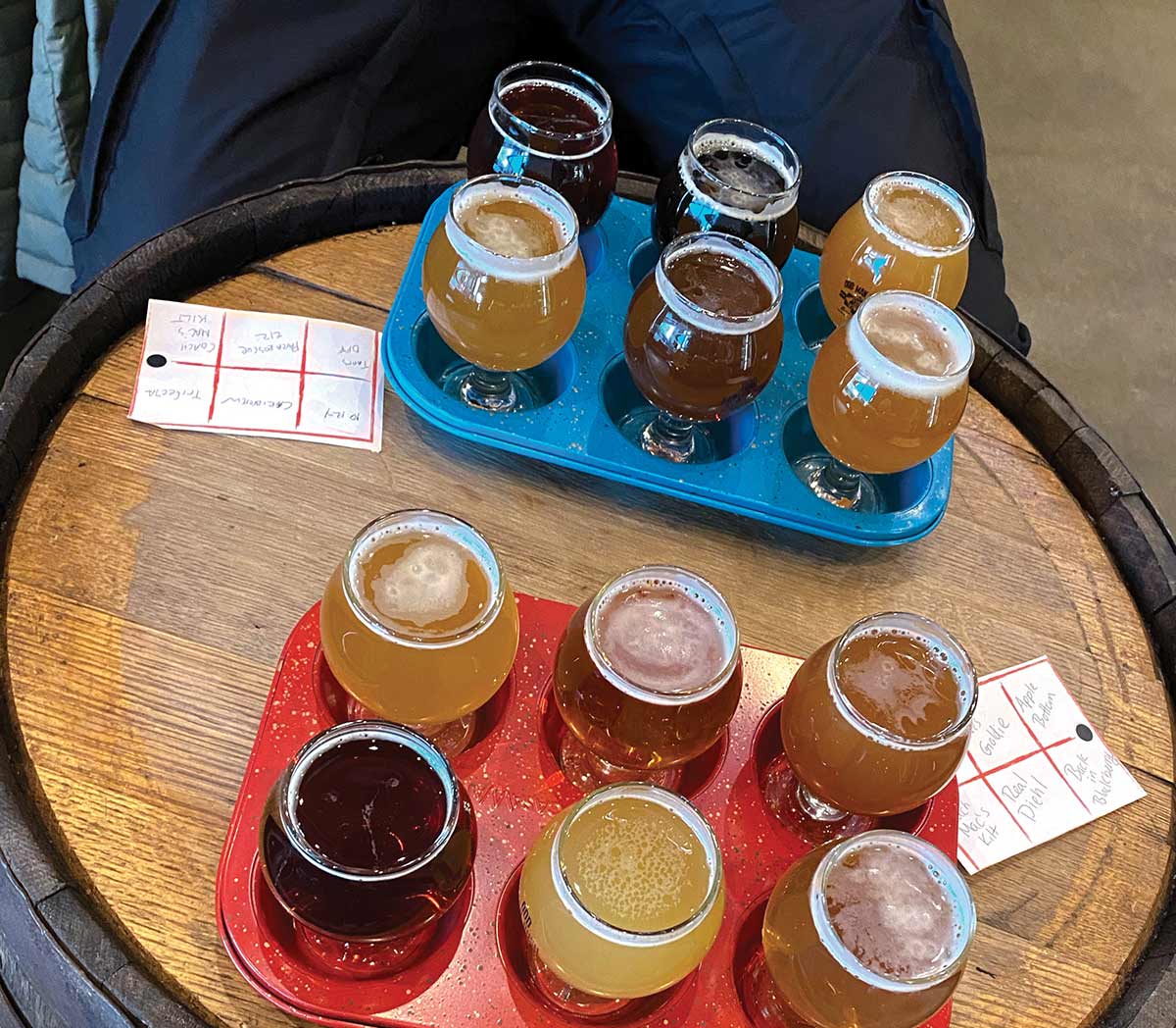 An overhead view of two beer flight samplers in red and blue holders on a wooden barrel table, featuring small glasses of various craft beers next to handwritten tasting notes.