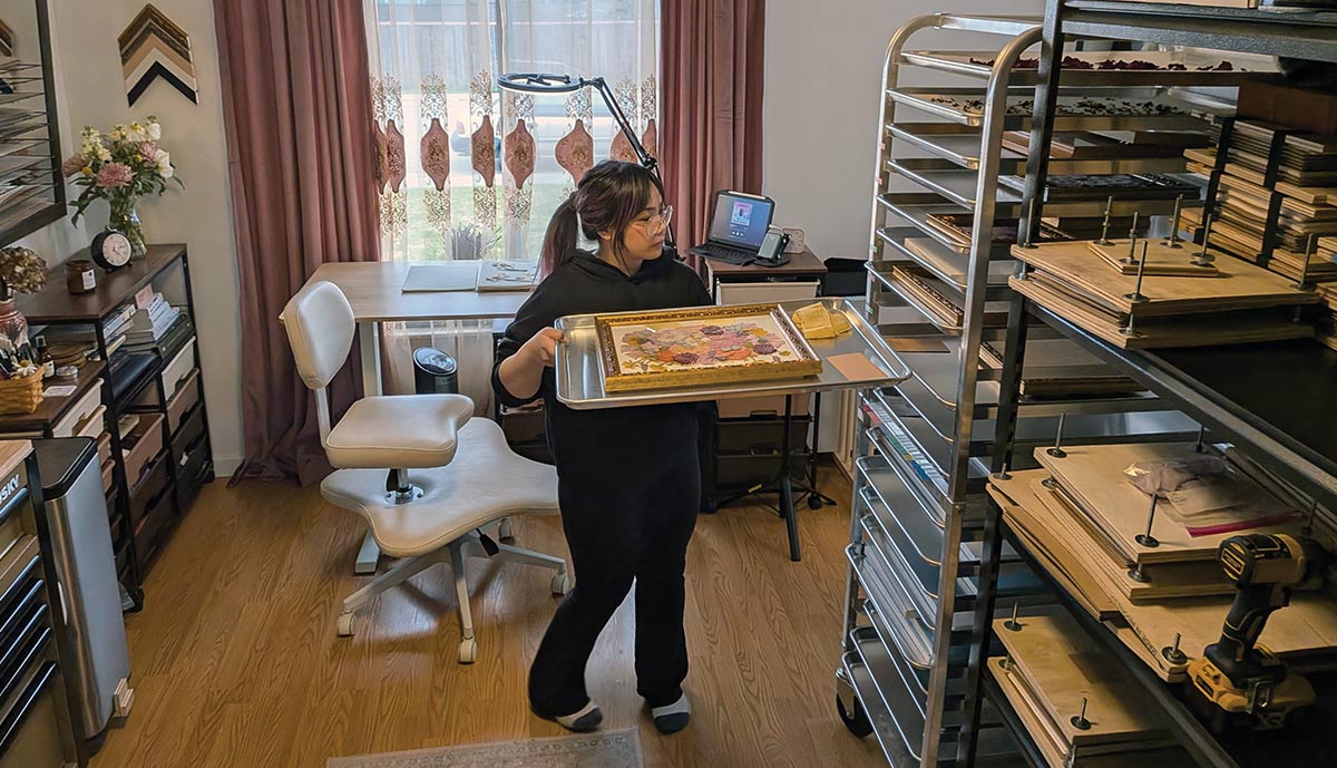 A woman in a home studio carries a metal tray with a framed floral artwork toward a tall stainless steel rolling rack, surrounded by shelves containing crafting tools and wooden presses.