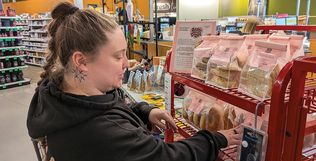 A woman with a neck tattoo stocks a red display rack in a grocery store with packaged "Wild About Dough" baked goods like cookies and bagels.