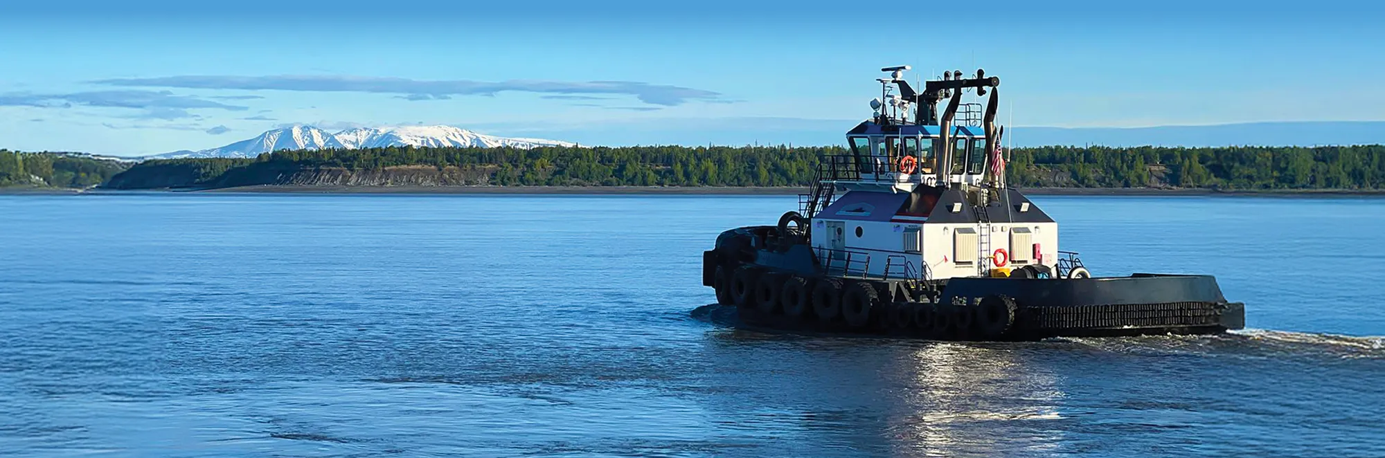 A tugboat travels across calm blue water, with a forested shoreline and distant snow-capped mountains visible under a clear blue sky.
