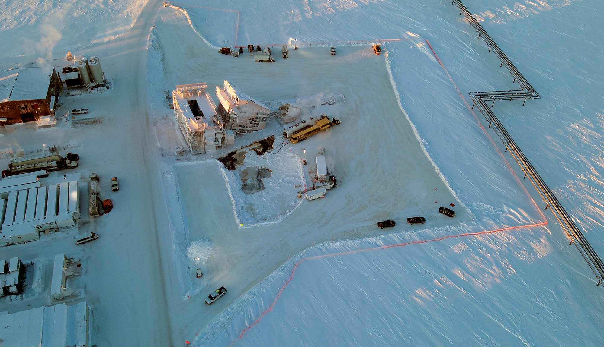 An aerial view shows an industrial facility and utility pipelines surrounded by a vast, snow-covered landscape