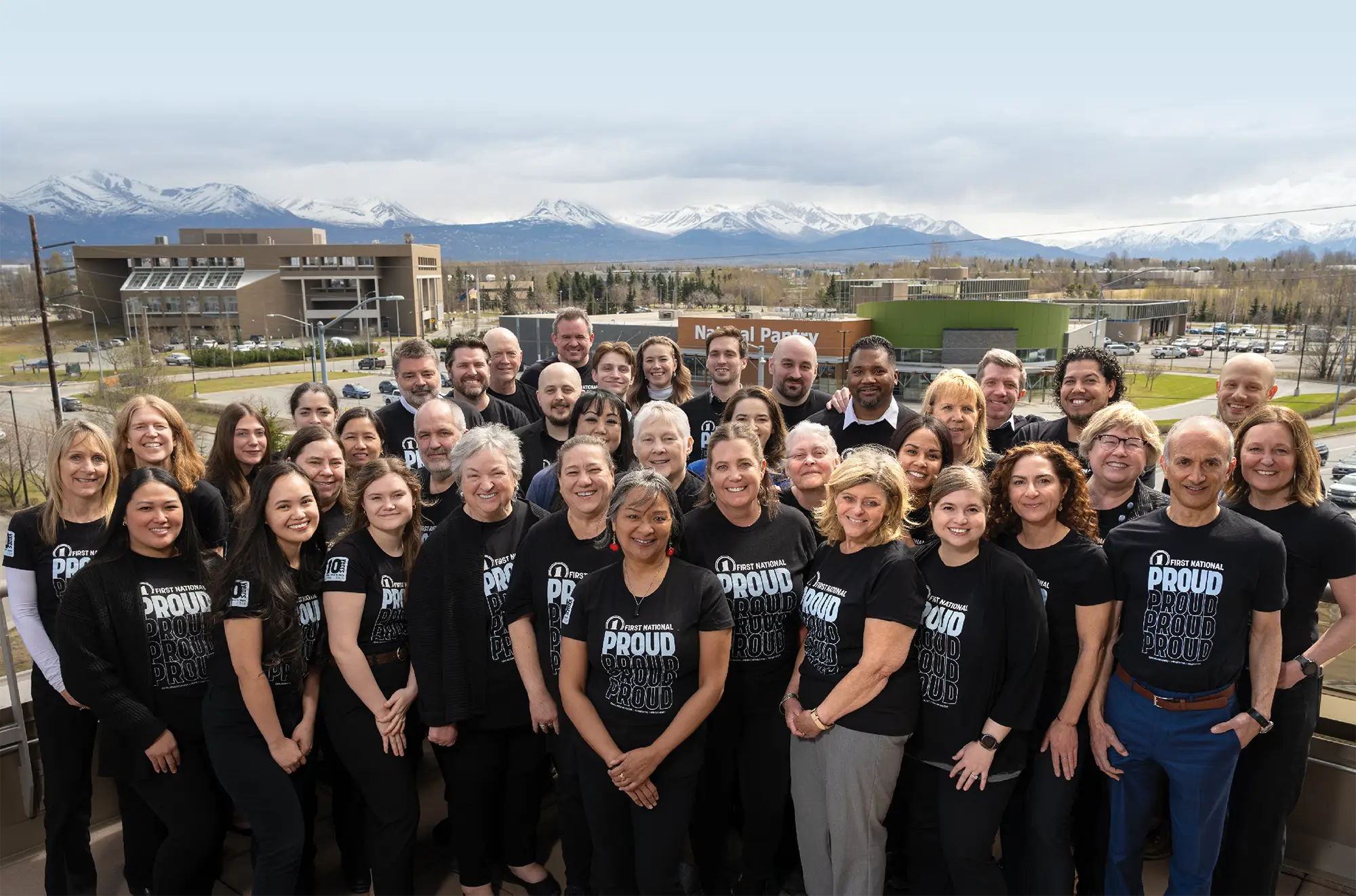 First National Bank Alaska employees wearing branded shirts pose together outdoors with Anchorage buildings and snow-covered mountains behind them.