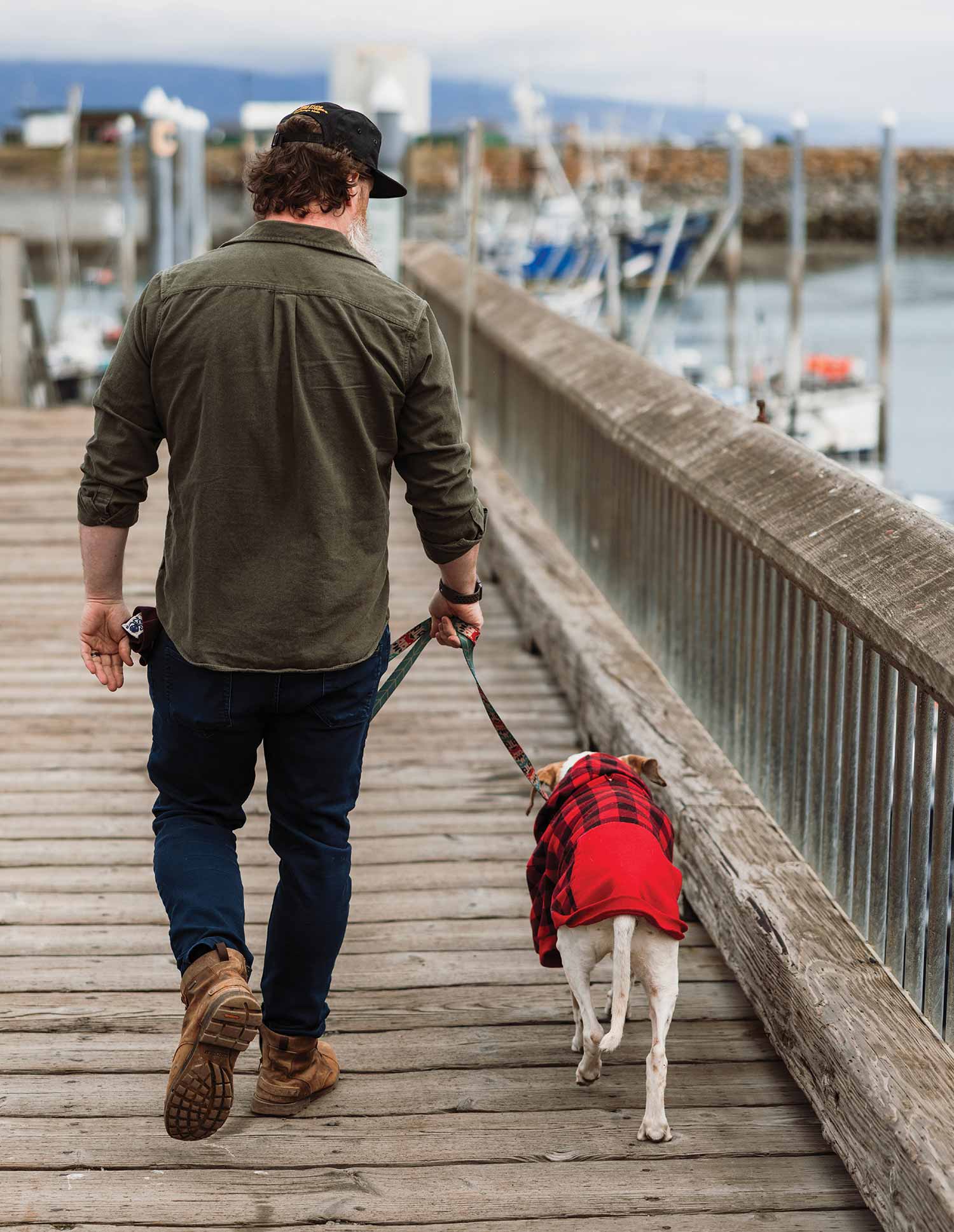 Lucas Elliott, a man viewed from behind walks down a wooden pier while walking a dog on a leash; The dog is wearing a red and black plaid pull-over sweater