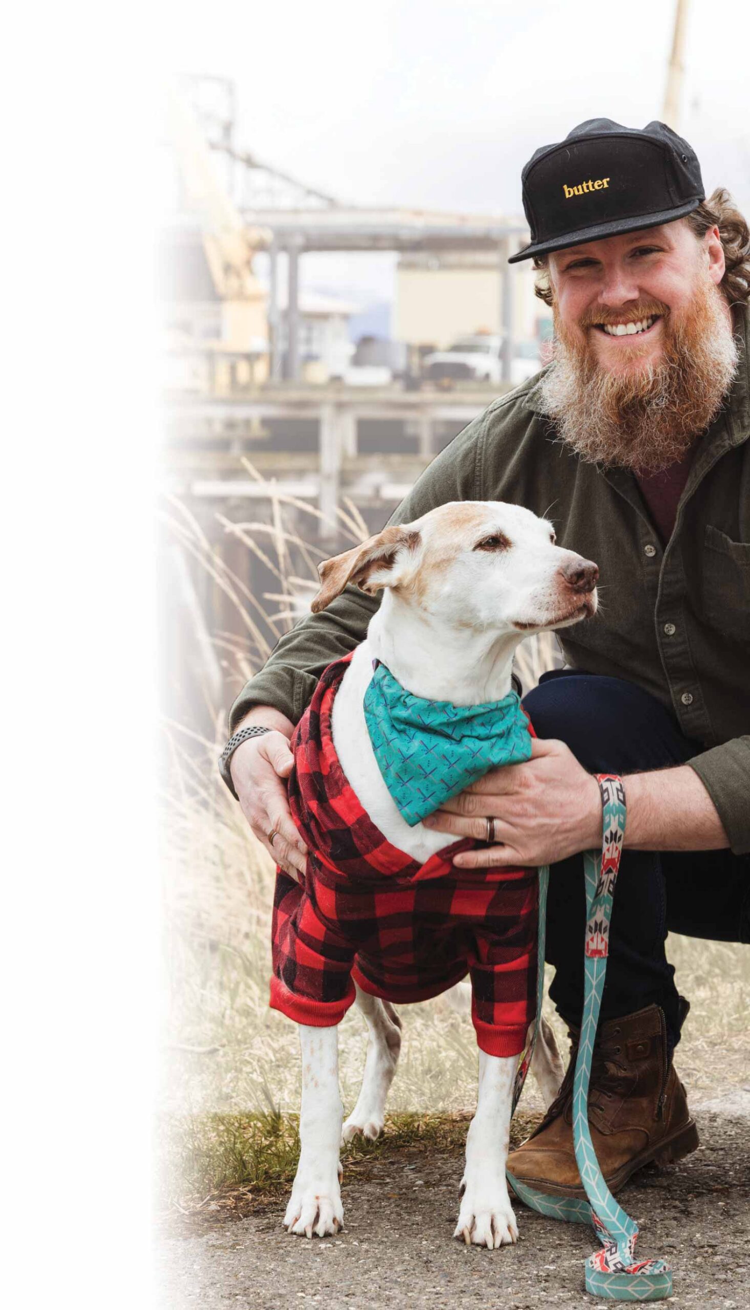 Lucas Elliott, a smiling, bearded man wearing a black hat with the word butter on it crouches down next to his dog outdoors in a wooden dock pier area