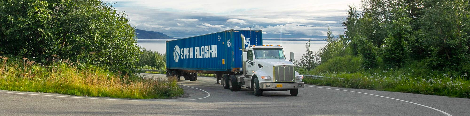 White semi-truck pulling a large blue shipping container with the Span Alaska logo drives along a wide, curving road surrounded by lush green trees