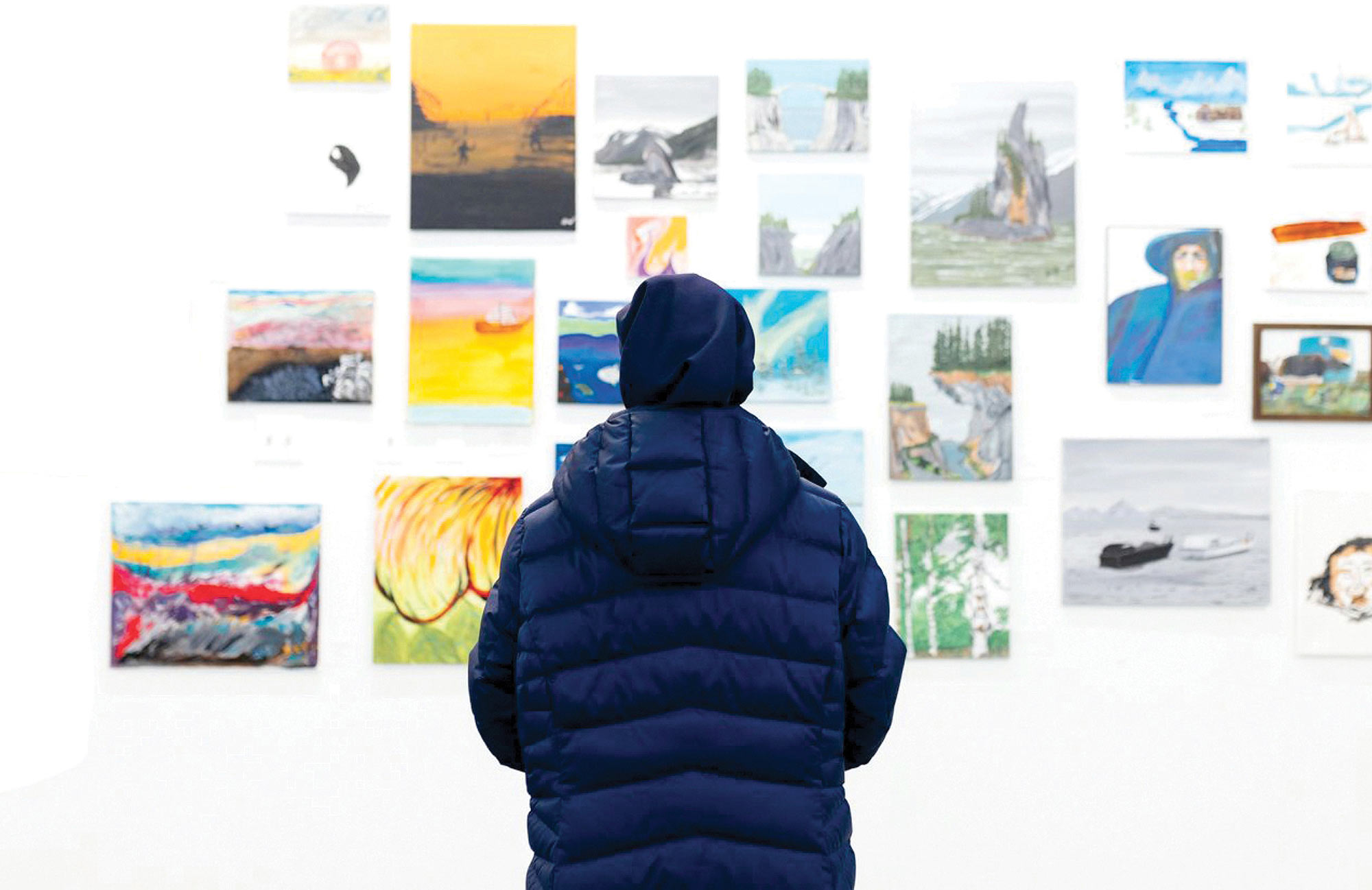 A person wearing a dark blue puffer jacket viewed from behind stands looking at a wall crowded with small, colorful landscape paintings.