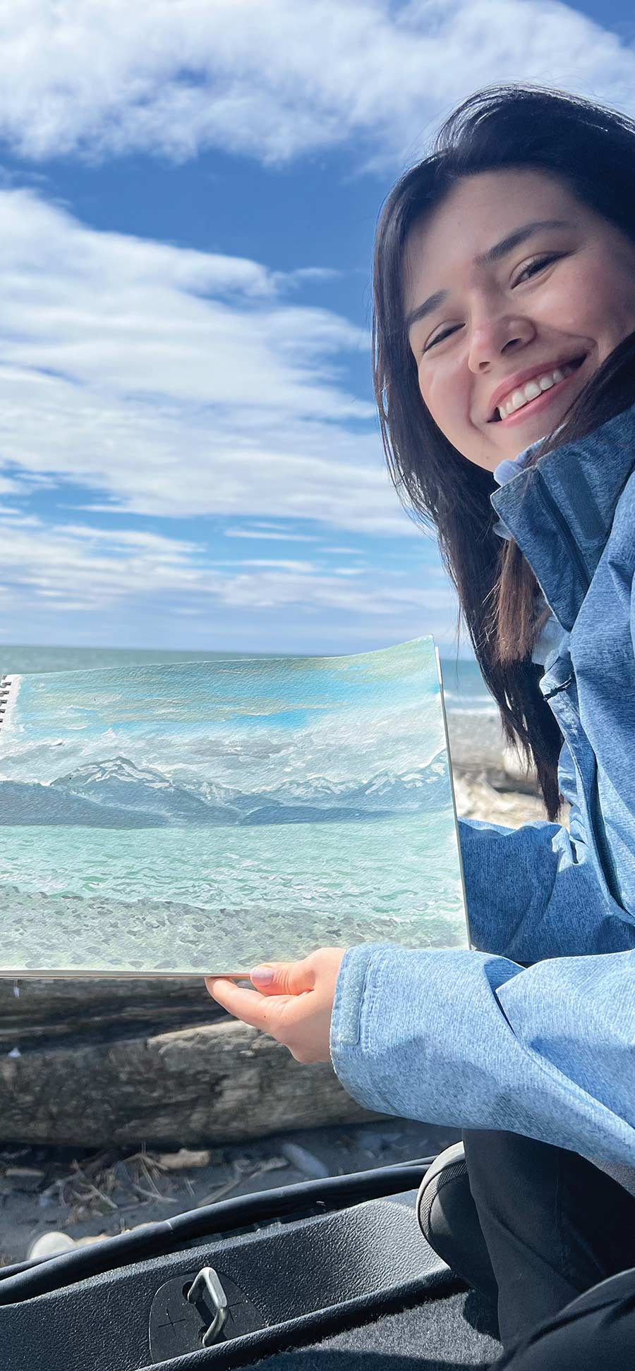 Valerie Jaimes in a light blue jacket poses outdoors while holding up a painting of a coastal mountain range under a cloudy sky.
