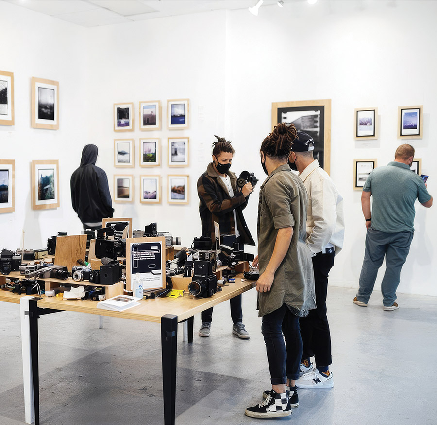 Visitors browse a gallery exhibition featuring a central table packed with vintage film and video cameras, with framed artwork displayed on the walls.