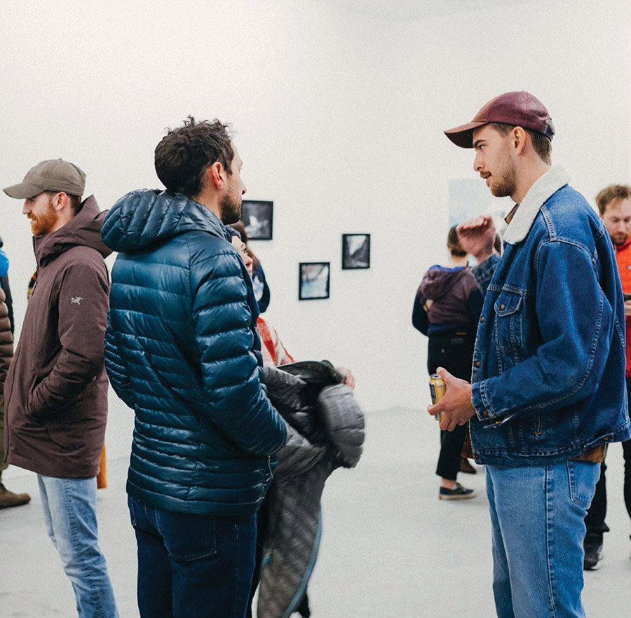 A group of people chat inside a brightly lit gallery space, with a man in a denim jacket and baseball cap talking to a person in a blue puffer jacket.
