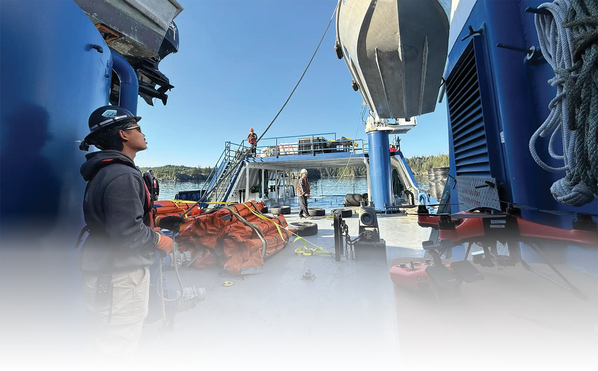Crew members work on the deck of a blue vessel under a clear blue sky, next to piled orange containment booms and a small red drone.