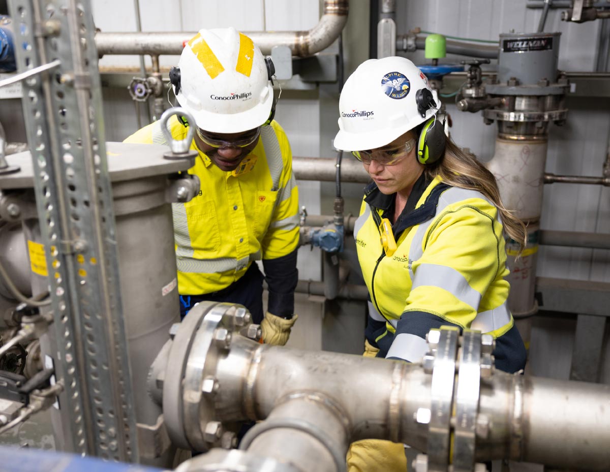 Two ConocoPhillips workers in hard hats and safety gear inspect a large industrial pipe network inside a facility.
