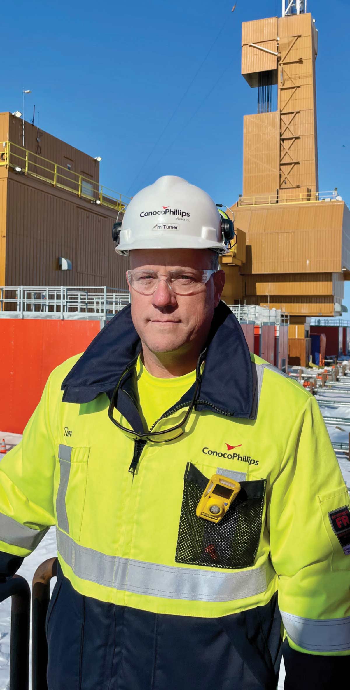 Portrait of a ConocoPhillips worker named Tim Turner wearing a hard hat and safety glasses, with an oil rig in the background.