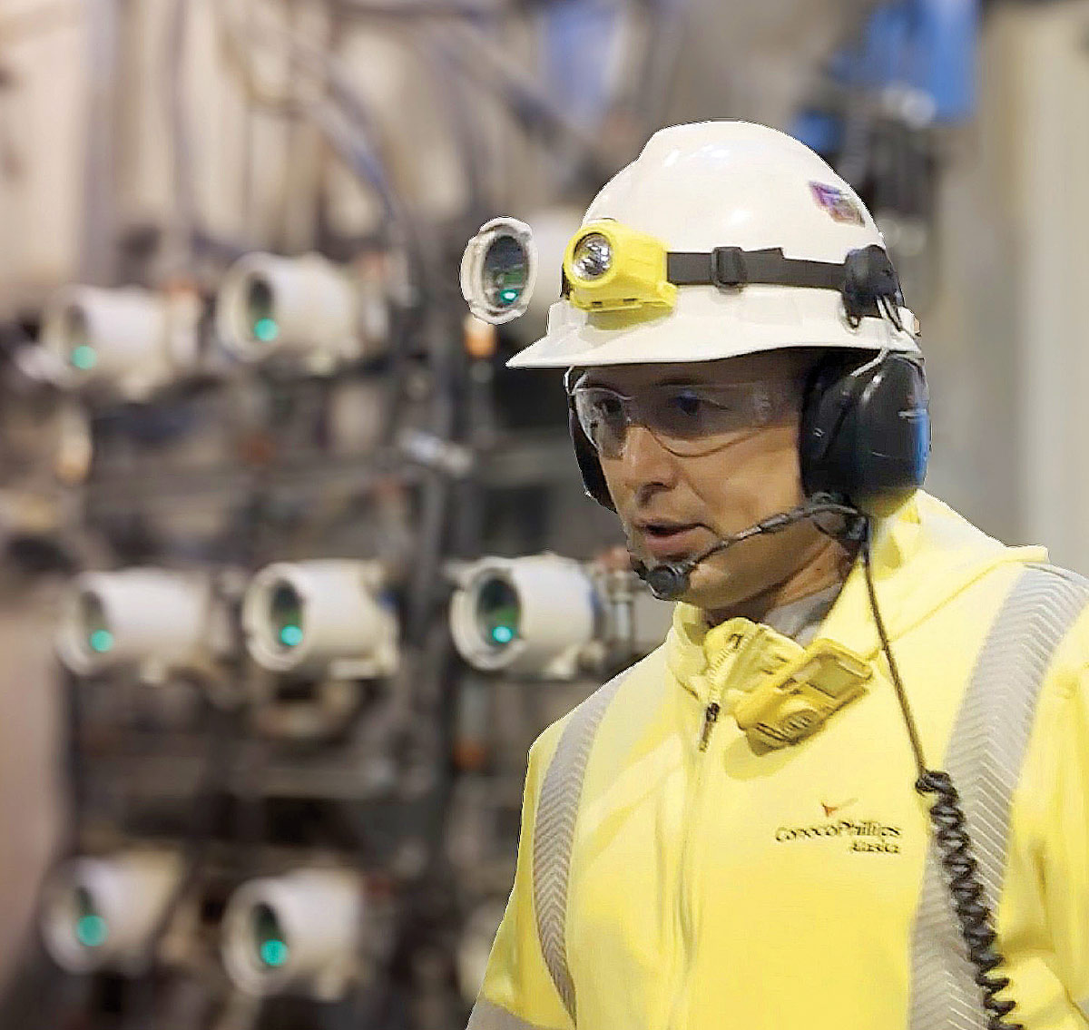 Close-up of a facility worker wearing a white hard hat with a headlamp, safety glasses, and a communication headset.