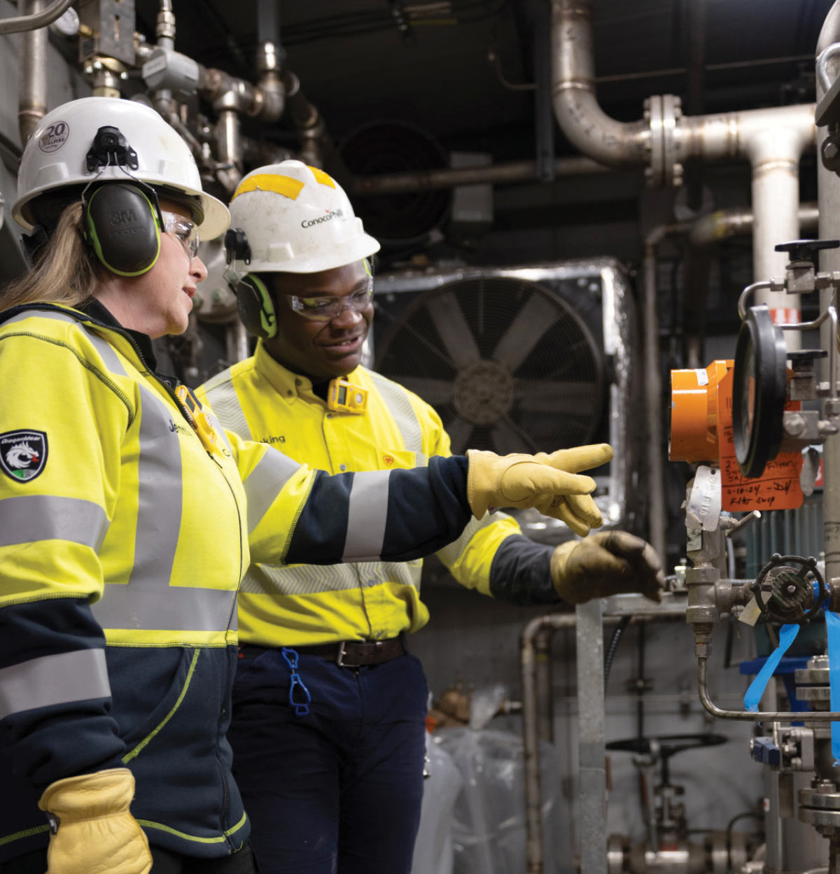 Two workers wearing bright safety jackets and hard hats point to and discuss equipment on an industrial piping system.