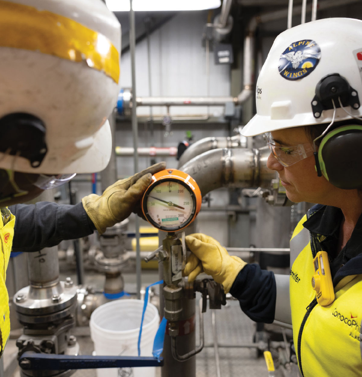 Close-up of two workers in protective gloves adjusting and checking the dial on a round pressure gauge attached to a pipe.