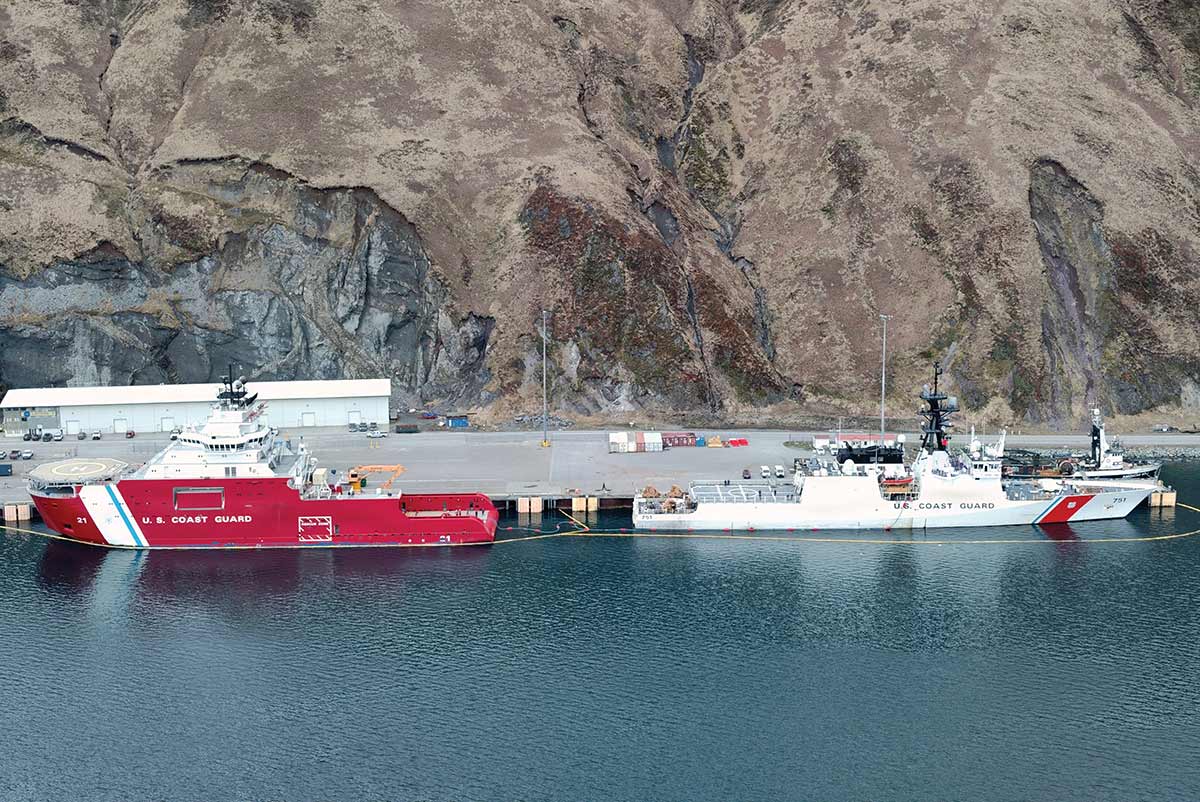 Two U.S. Coast Guard vessels, a red icebreaker and a white cutter, are docked at a harbor pier in front of a steep, barren mountainside.