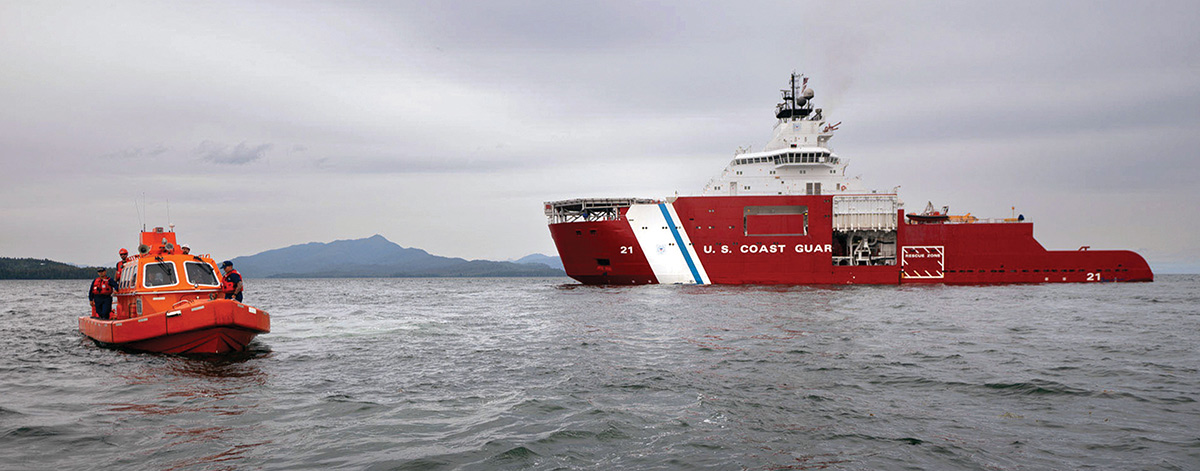 A small orange Coast Guard rescue boat with crew members sails in the foreground, with a large red and white U.S. Coast Guard vessel in the background on open water with distant mountains.