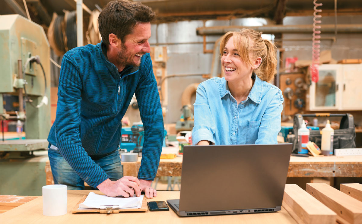 A smiling man and woman look at each other while reviewing data on a laptop in a woodworking workshop.