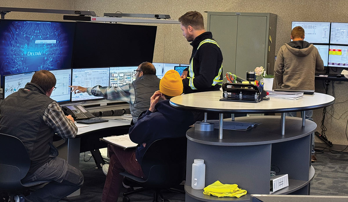 Operators work together in an industrial control room, monitoring technical data and schematics on a wall of computer screens while one man points out information.