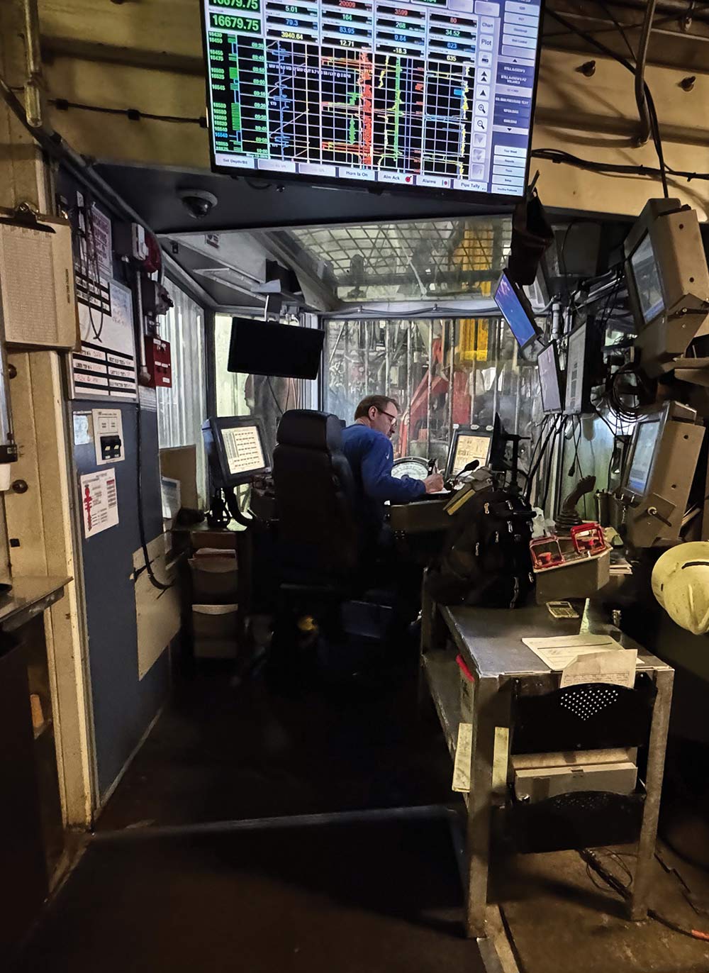 An operator sits in a control chair inside a drilling rig cabin, looking through a window at machinery while monitoring technical data and graphs on multiple screens.