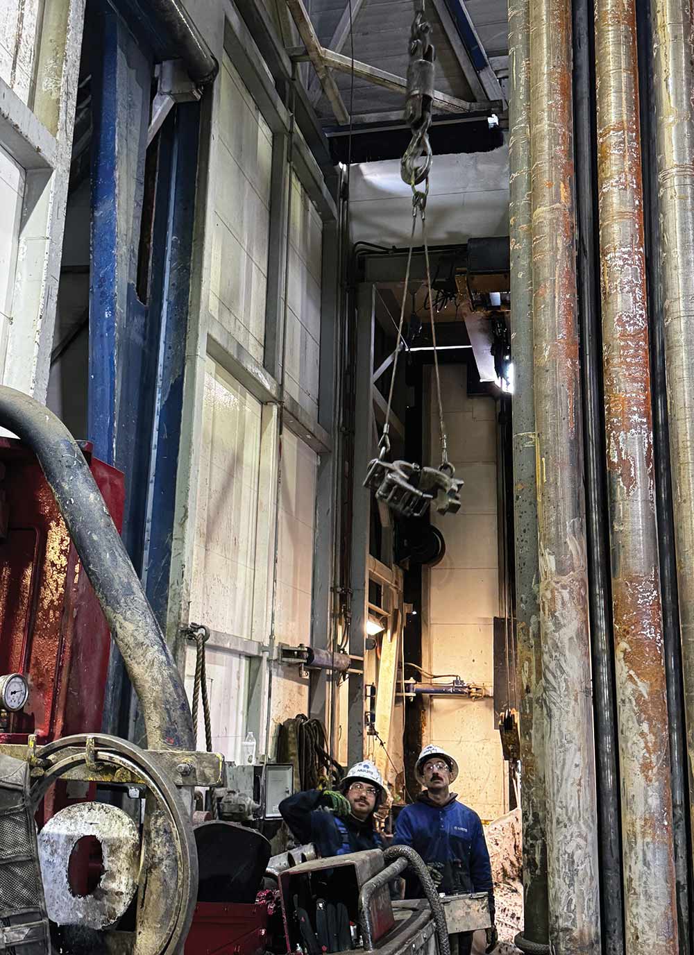 Two rig workers wearing hard hats look upward at industrial equipment suspended from a hoist inside a drilling tower next to large vertical pipes.