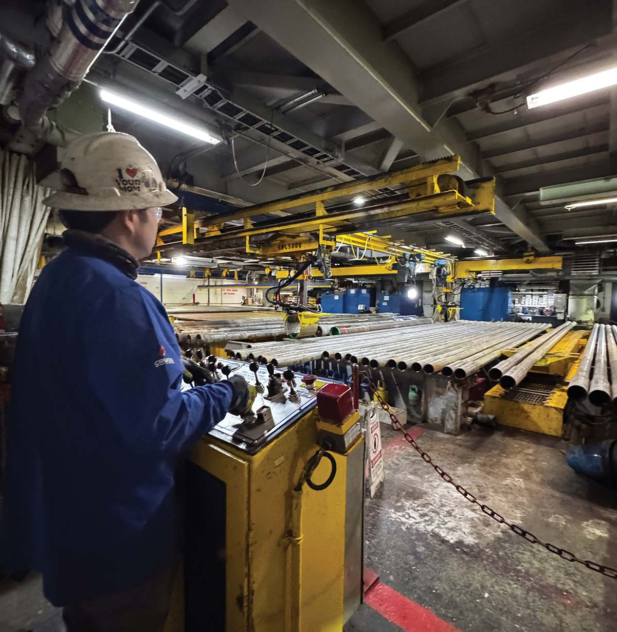 A worker wearing a hard hat operates a yellow control panel with levers inside an industrial facility, overlooking large racks of long metal pipes.