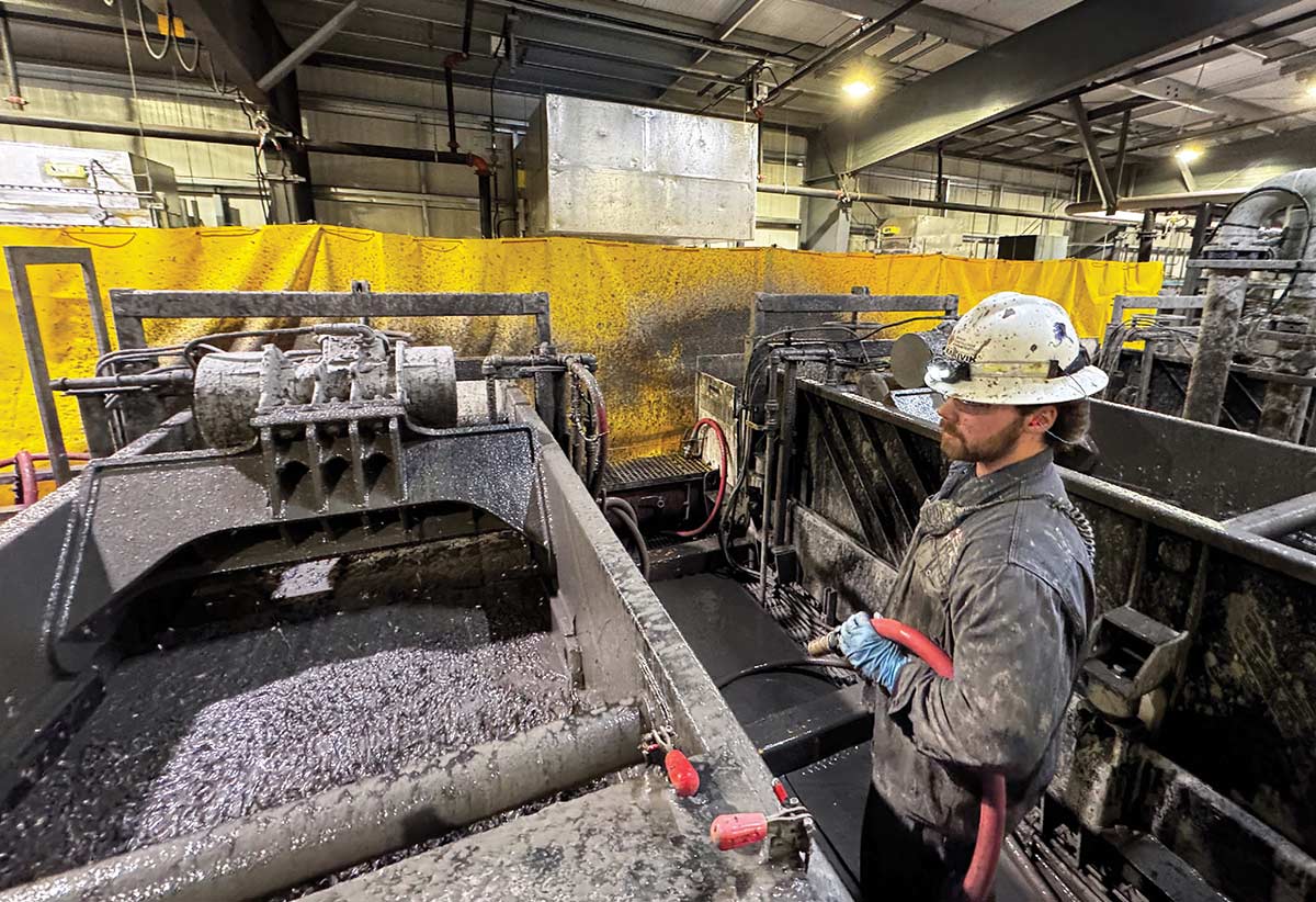 A worker in a mud-splattered hard hat and jacket holds a red hose while standing next to large industrial machinery processing drilling fluid inside a facility.