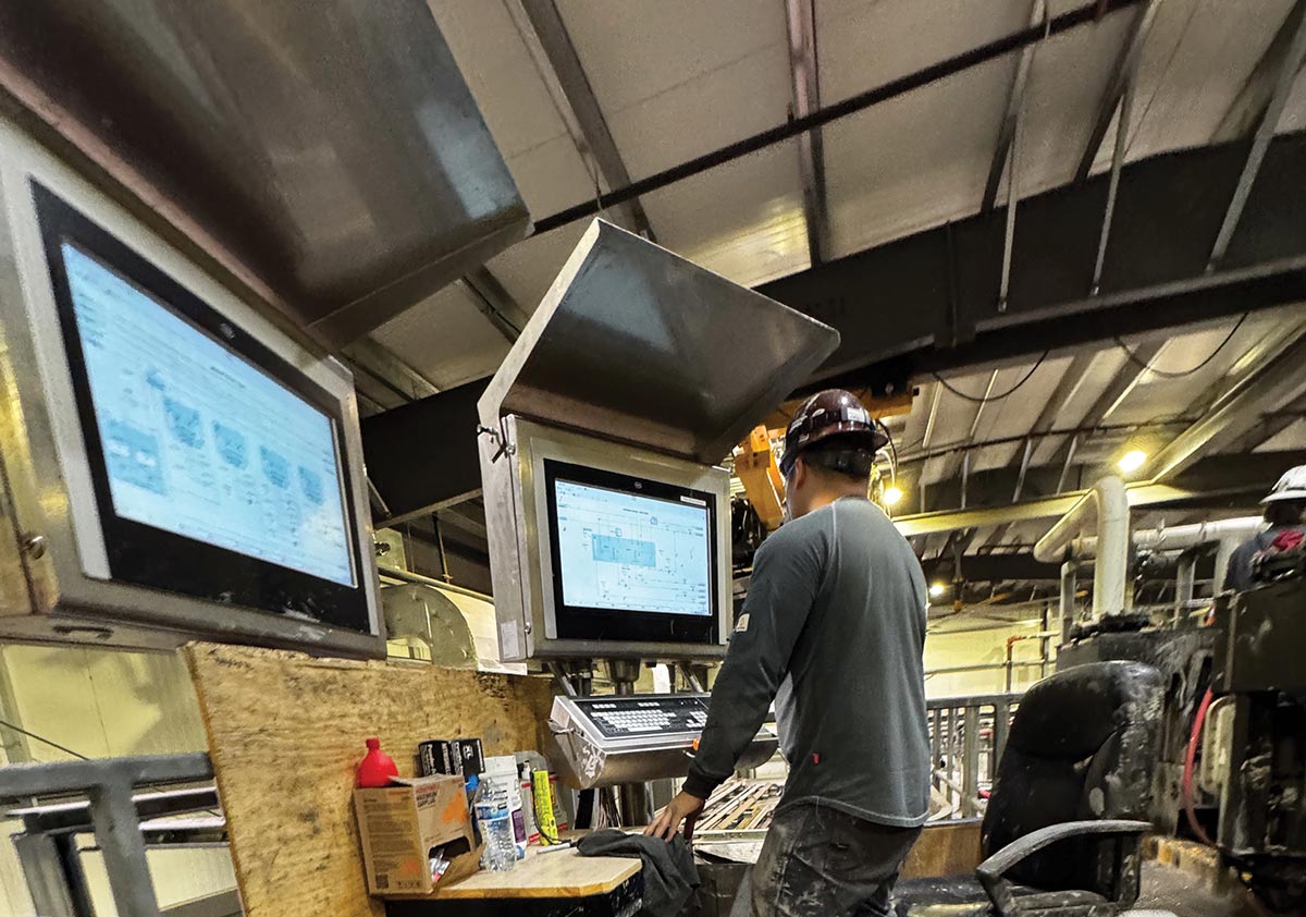 A worker wearing a hard hat stands at an industrial workstation inside a facility, monitoring data and technical schematics on computer screens.