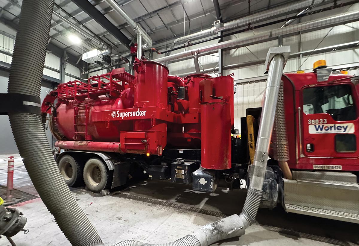 A large red Worley vacuum truck with "Supersucker" written on the side is parked inside an industrial facility with overhead pipes and ventilation hoses.