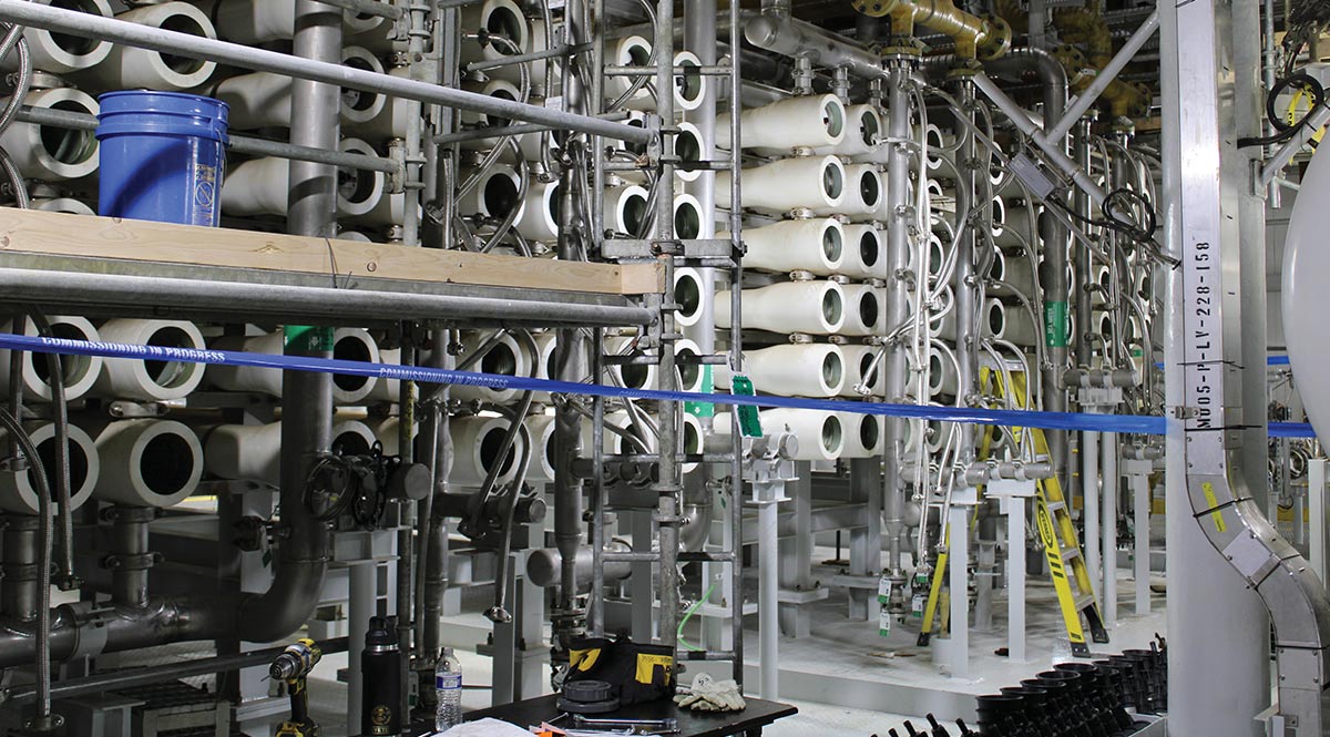 Rows of white industrial cylindrical vessels and complex metal piping inside a facility, with blue tape reading "COMMISSIONING IN PROGRESS" stretched across scaffolding in the foreground.