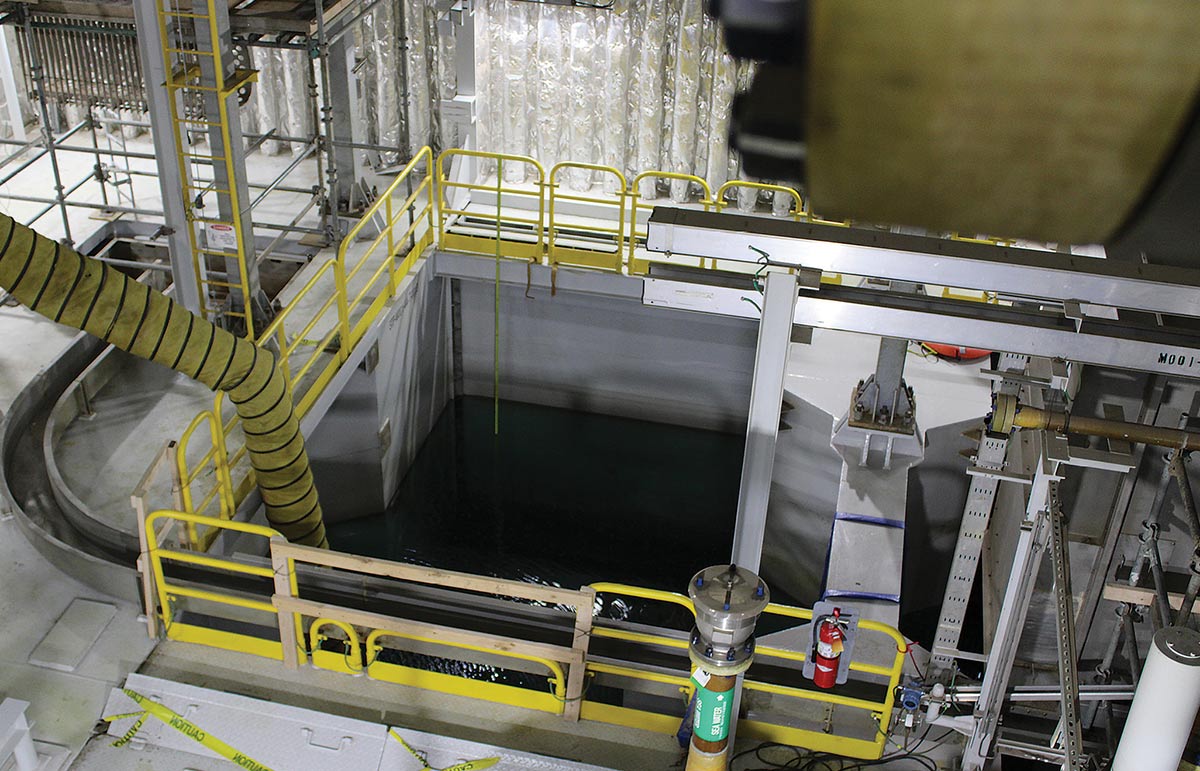 An elevated view of a large concrete basin filled with dark water inside an industrial facility, surrounded by yellow safety railings, walkways, and piping.
