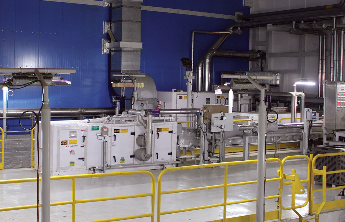Interior of an industrial facility featuring white machinery units and complex metal piping against a bright blue wall, viewed from behind yellow safety railings.