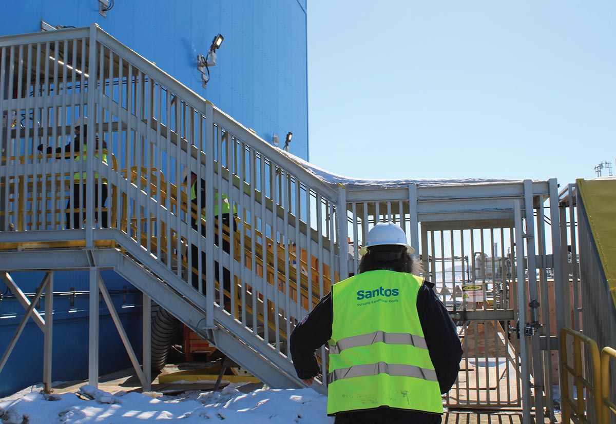A worker in a yellow Santos safety vest and hard hat looks toward an outdoor metal staircase where other workers climb the side of a large blue industrial building.