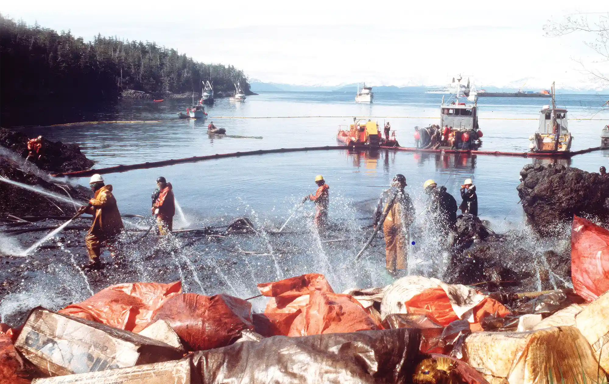 Cleanup workers in protective gear spray water hoses to wash a rocky shoreline. Containment booms and response boats are visible in the water in the background.
