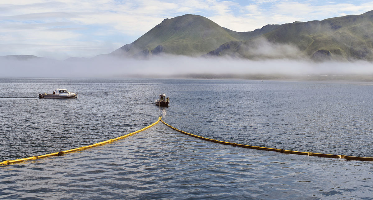 Two small boats deploy a yellow containment boom in calm coastal waters, with low-lying fog and grassy mountains in the background.